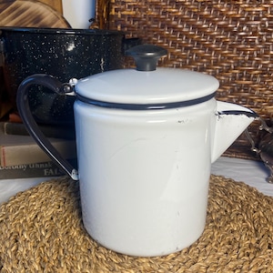 May include: White enamel coffee pot with a black handle and lid. The pot has a classic design with a spout for pouring. A black speckled pot is visible in the background. The coffee pot is sitting on a woven mat.