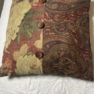 May include: Square decorative pillow with a floral and paisley pattern. The left side features large cream-colored flowers on a burgundy background. The right side has a paisley design in shades of burgundy, green, and brown. Three brown buttons are sewn on the seam.