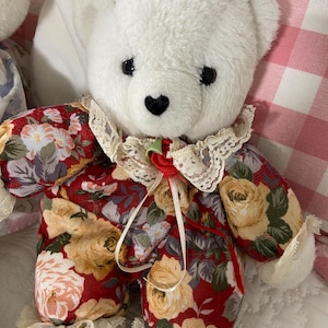 May include: A white plush teddy bear with a heart-shaped nose and black eyes. The bear wears a red floral print dress with a lace collar and cuffs. A red ribbon and rose accent the neckline. The background includes a pink and white checkered pattern.