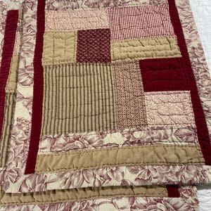 May include: Two quilted placemats with a patchwork design. The placemats feature a mix of patterns, including floral, striped, and solid fabrics in shades of red, beige, and cream. The edges are trimmed with a deep red fabric.