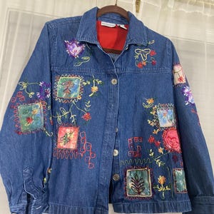 May include: A blue denim jacket featuring a collar, long sleeves with buttoned cuffs, and a front button closure. The jacket is decorated with various colorful embroidered patches, floral motifs, and stitched patterns in red, green, and purple.