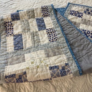 May include: A patchwork quilt in shades of blue, gray, and white. The quilt features a grid pattern with squares of solid colors and patterned fabrics. The edges are finished with a blue binding.