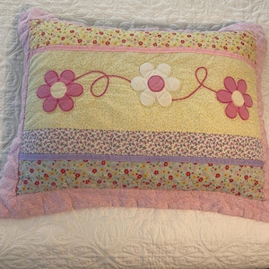 May include: A rectangular, quilted pillow with a floral design. The pillow features a light yellow center panel with three appliqued flowers connected by a pink stem. The border and backing are a soft pink.