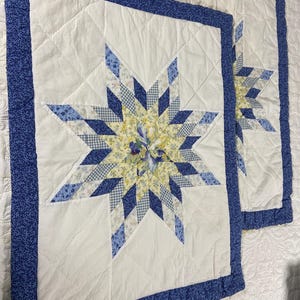 May include: Two quilted wall hangings with a star design. The stars feature blue and white patterned fabrics, with a yellow floral center. The borders are a solid blue with a floral pattern. The background is white.
