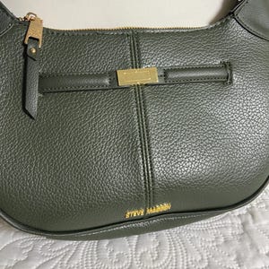 May include: A dark olive green hobo bag with a pebbled texture, featuring a gold-tone zipper closure, a matching zipper pull, and a horizontal decorative gold-tone metal bar. The brand name STEVE MADDEN is printed in gold lettering at the bottom.
