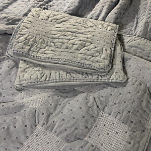 May include: A light blue quilted bedspread and two matching pillow shams. The bedspread features a diamond pattern with small floral accents. The pillow shams have a textured, stitched design. The bedding is made of a soft, possibly cotton, material.