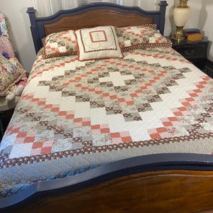 May include: A bed featuring a quilt with a patchwork design in shades of coral, brown, and cream, arranged in a diamond pattern. The bed includes two matching rectangular pillows and a central square pillow with a floral embroidery design.
