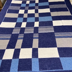 May include: A navy blue and white checkered blanket with light blue and gray accents. The blanket has a dark blue border and a geometric pattern. The blanket is made of a soft material and is suitable for a bed or couch.
