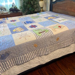 May include: A nautical-themed quilt with a patchwork design. The quilt features squares in shades of blue and white, with embroidered images of sailboats, compasses, and maps. The quilt has a striped border and a white bed skirt.