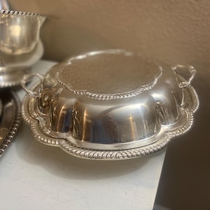 May include: A round, silver-toned serving dish with a domed lid, featuring a scalloped edge and a decorative rope-style border. The piece includes two small, curved handles on opposite sides and a polished, reflective metallic finish.