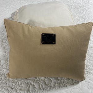 May include: A rectangular tan pillow with a black leather patch in the center. The pillow is on a white quilted bedspread, with a white round pillow in the background.