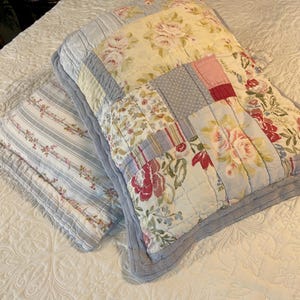 May include: Two decorative pillows on a white bedspread. One pillow features a patchwork design with floral patterns in pink, red, and blue. The other pillow has a striped pattern with floral accents. Both pillows have a quilted appearance.