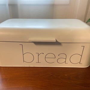 May include: Off-white rectangular bread box with a curved lid and a handle. The word "bread" is printed in black, stylized font on the front of the box. The bread box is sitting on a wooden surface.