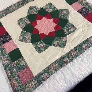 May include: A square quilted wall hanging featuring a central star motif in shades of pink, dark green, and floral patterns. The design is surrounded by a cream-colored border with curved stitching and an outer frame of alternating floral and solid squares.