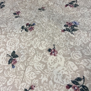 May include: Beige fabric with a floral pattern. The design features large, off-white flowers and leaves, with smaller clusters of pink and purple flowers scattered throughout. The fabric has a soft, textured appearance.
