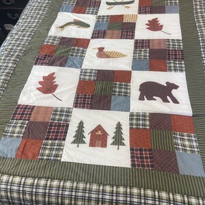 May include: A patchwork quilt featuring a central grid of white squares with appliquéd motifs including a canoe, fish, leaf, duck, bear, and cabin with trees. The quilt is bordered by green striped fabric and a plaid outer edge in earth tones.