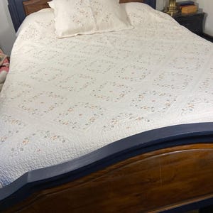 May include: A bed frame with a dark blue painted wooden headboard and footboard featuring natural wood panels. The bed is covered with a white quilt featuring a repeating diamond pattern with embroidered floral motifs and a matching pillow.
