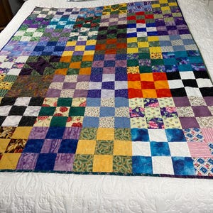 May include: A handmade quilt with a patchwork design. The quilt features a checkerboard pattern with squares in various colors, including blue, green, yellow, red, and purple. The quilt is placed on a white bedspread.