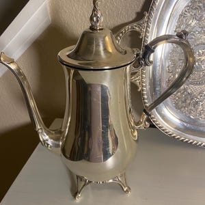 May include: A polished silver-toned coffee pot featuring a tall, slender body, a curved spout, and a decorative finial on the lid. The pot stands on three ornate feet and includes a curved handle with black accents, positioned next to a silver tray.