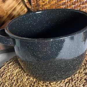 May include: A black speckled enamelware pot with two handles. The pot is sitting on a woven placemat. The pot has a shiny finish and a wide opening. The background includes a wooden bowl and a wicker basket.
