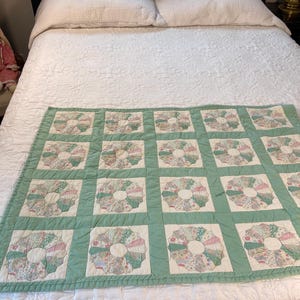 May include: A rectangular quilt featuring a repeating pattern of circular floral fabric blocks in shades of pink, green, and white. Each circular block is framed by a white square border and separated by a solid light green fabric grid.