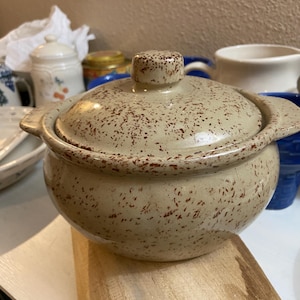 May include: A round ceramic casserole dish with a matching lid, featuring a light beige base color with a speckled brown pattern throughout. The pot has two small side handles and a central knob on the lid, with a smooth, glazed finish.
