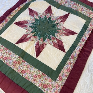 May include: A quilted fabric piece featuring a central star pattern in shades of dark green and burgundy. The star is surrounded by cream-colored squares, a floral print border with roses, and a solid burgundy outer border with stitching.