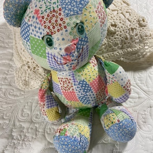 May include: A handmade patchwork teddy bear with a variety of colorful floral and geometric patterned fabric squares. The bear has blue ears, green eyes, and a pink nose. The bear is sitting on a white, quilted surface.
