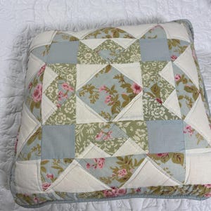 May include: Square decorative pillow with a patchwork design. The pillow features floral patterns in pink, green, and blue, with white and cream-colored accents. The pillow is placed on a white quilted surface.