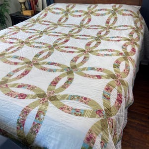 May include: A bed quilt featuring a white background with a repeating double wedding ring pattern. The interlocking rings are constructed from fabric strips in shades of sage green, soft pink, and cream, decorated with floral and striped prints.