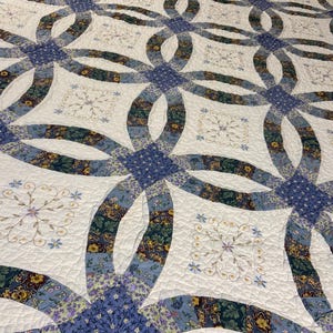 May include: A double wedding ring quilt featuring a white background with intricate blue, green, and yellow floral patterned fabric segments. The quilt includes embroidered floral motifs within the white squares and a quilted texture throughout.