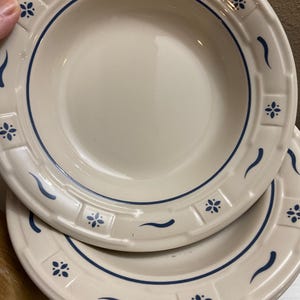 May include: A set of two cream-colored ceramic bowls featuring a wide rim with a raised grid pattern. The design includes dark blue painted accents consisting of small floral motifs and curved lines spaced evenly around the border of each bowl.