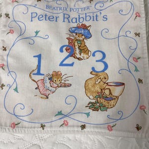 May include: A white fabric square featuring illustrations of Peter Rabbit characters and the numbers 1, 2, and 3. The design includes a blue border with floral accents and the text "Beatrix Potter Peter Rabbit's".