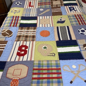 May include: A patchwork quilt featuring sports-themed squares. Designs include a football, basketball, baseball, soccer ball, and a football helmet. The quilt has a blue, green, and brown color scheme with red and white accents.
