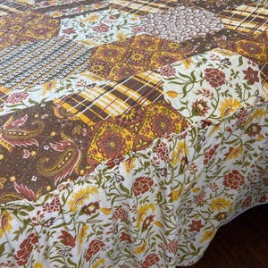 May include: A patchwork quilt featuring a hexagonal pattern with various fabric prints in shades of brown, mustard yellow, orange, and white. The quilt includes floral, paisley, and plaid designs, finished with a ruffled floral edge trim.