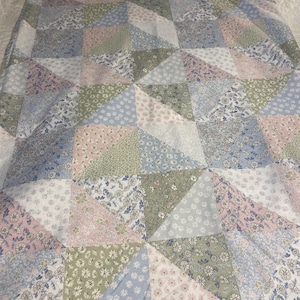 May include: A patchwork quilt featuring a repeating triangular pattern composed of various floral fabrics in soft shades of sage green, dusty pink, light blue, and white. The textile displays a consistent geometric arrangement of small-scale botanical prints.