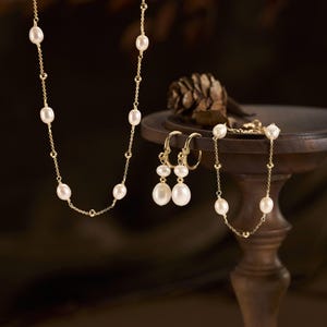 May include: A gold chain necklace featuring spaced white oval pearls, a pair of gold hoop earrings with dangling white oval pearls, and a gold chain bracelet with spaced white oval pearls. The text PastelCharmCoStore is visible in the corner.