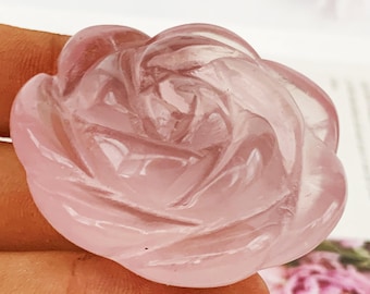 May include: A rose-shaped gemstone, carved in a delicate pink colour. The gemstone is held by a person's fingers. The rose is intricately detailed, with each petal carefully sculpted. The overall effect is delicate and elegant, suggesting a piece of jewellery or a decorative item.