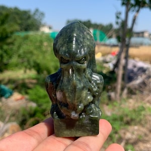 Può includere: Una scultura in pietra verde scuro raffigurante una creatura con il volto tentacolare e la testa a punta. L'opera presenta occhi infossati e una superficie strutturata, poggia su una base rettangolare e mostra variazioni minerali naturali.