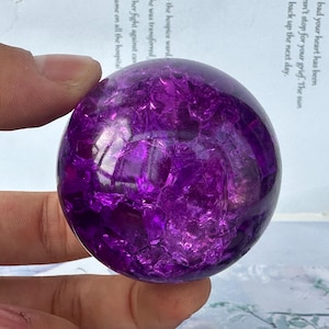 Puede incluir: Una vibrante bola de cristal de amatista esférica. La esfera pulida es de color púrpura intenso con estructuras cristalinas internas. La superficie refleja la luz, creando un efecto brillante. El cristal se sostiene en una mano.