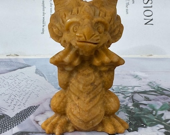 May include: A carved figurine of a mythical creature in a light brown color. The figure has a lion-like face with large ears and is holding its hands up to its face. The figurine is standing upright with its legs spread apart.