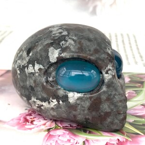May include: A grey alien head sculpture with large, oval, blue eyes. The sculpture has a textured surface with silver accents. The alien head is a unique decorative item.