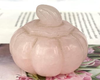 May include: A light pink rose quartz pumpkin carving. The pumpkin has a rounded body with vertical grooves and a curved stem. The figurine is displayed on a floral background, suggesting a decorative or seasonal item.