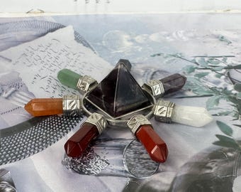 May include: A crystal energy generator with a dark pyramid at the centre, encircled by seven colourful gemstone wands. The wands are in shades of red, orange, green, purple, and white, set in silver-coloured metal. The background is a patterned surface.
