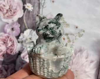 May include: A carved stone cat figurine in a basket. The cat is white with green veining, and the basket has a woven texture. The background features soft pink and white flowers.