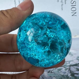 May include: A clear, blue sphere with a cracked, textured surface. The sphere is held in a hand and appears to be made of glass or crystal. The color is a vibrant turquoise, and the cracks create a unique pattern.