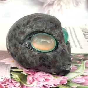 May include: A grey stone alien head sculpture with oval, iridescent eyes. The eyes are a mix of orange, green, and blue. The sculpture is set against a floral background.