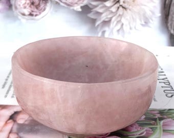 May include: A rose quartz bowl with a smooth, polished surface. The bowl is a soft pink color and has a rounded shape. The background features blurred flowers.