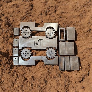 K&ouml;nnte beinhalten: Unfertige Metallteile, angeordnet auf einer braunen Oberfl&auml;che. Darunter zwei jeepartige Formen mit zahnradf&ouml;rmigen R&auml;dern, ein rechteckiges Teil mit der Gravur "WT" und verschiedene rechteckige und quadratische Komponenten. Das Thema deutet auf ein DIY-Projekt oder Metallkunst hin.