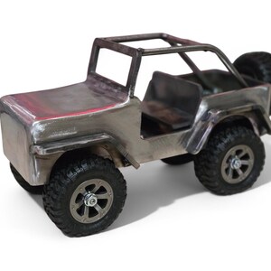 K&ouml;nnte beinhalten: Ein handgefertigter Metall-Spielzeug-Jeep mit &Uuml;berrollk&auml;fig und Ersatzreifen. Der silberfarbene Jeep hat schwarze Reifen und einen detaillierten Innenraum. Der Spielzeug-Jeep ist eine Miniatur-Nachbildung eines klassischen Gel&auml;ndewagens.